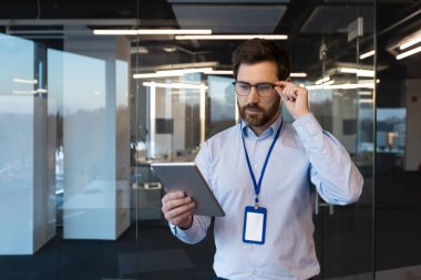 Şaşkın ve ciddi genç adam uzman ve müteahhit, ofis merkezinin binasında, bir gömlek ve rozetle duruyor, mühendislik konferansında yer alıyor, bardakları tutuyor ve