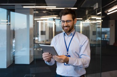 Gülümseyen genç adam, işçi ve uzman pencerenin yanında duruyor, elinde bir tablet tutuyor ve kullanıyor. Video çağrısı üzerinden çevrimiçi görüşmeler, veri ve projelerle çalışır.