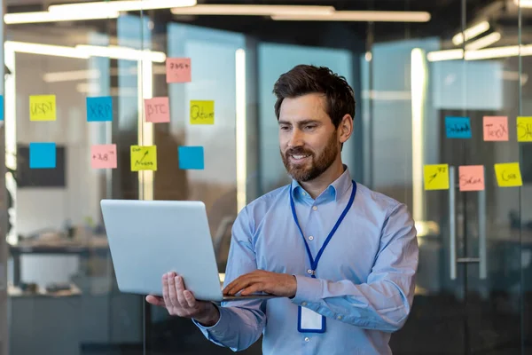 Gülümseyen genç iş adamı, programcı ve müteahhit bir iş merkezinin arka planında notlarla oturuyor ve konsantre bir dizüstü bilgisayar kullanıyor..