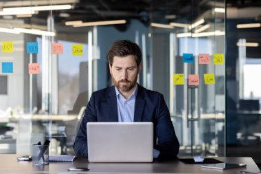 İş yerinde ciddi yoğunlaşmış finans işadamı elektronik mali rapor hazırlıyor, iş elbisesi giymiş olgun bir adam dizüstü bilgisayarda klavye yazıyor, içeriden rapor okuyor..