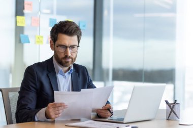 Ciddi düşünen bir iş adamı evrak işlerinde tecrübeli bir patron raporlar ve finansal belgelerle çalışıyor, finansör ofis içinde dizüstü bilgisayarlı, okuma sözleşmeli, düşünceli bir şekilde kontrol ediyor..