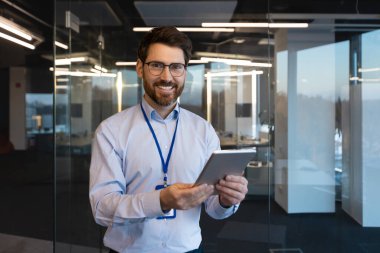 Olgun deneyimli programcı sistem yöneticisi portresi, elinde tablet bilgisayarla gülümseyen ve kameraya bakan adam, iş yerindeki yeni yazılımı test eden olgun işçi..