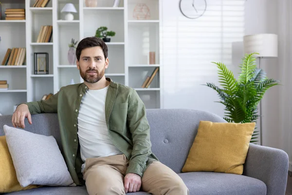Confident brunette man in casual outfit leaning on back of sofa with bend arm in stylish interior. Handsome apartment owner feeling satisfied with investment in new living room design at home.