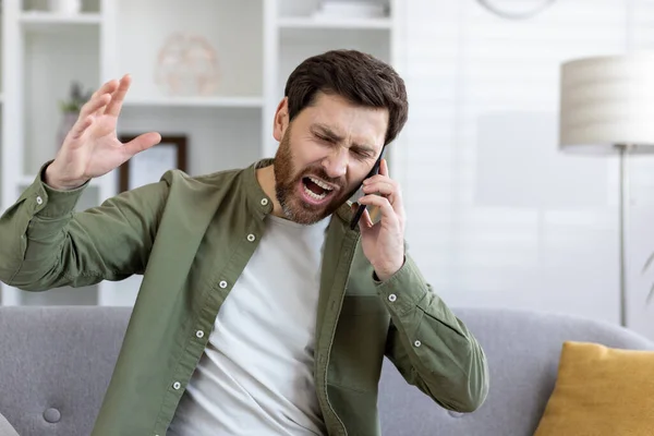 Mad unshaved man talking by cellphone and shouting angrily while raising hand in living room. Emotional guy having stressful conversation with technical support agent and enraging with bad service.