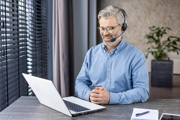 Kulaklıklı, mikrofonlu, elleri masanın üzerinde katlanmış, taşınabilir bilgisayar ekranına bakan memnun bir girişimci. Memnuniyet verici patron, çalışanların evde daha iyi sonuçlar aldığına dair raporları dinliyor..