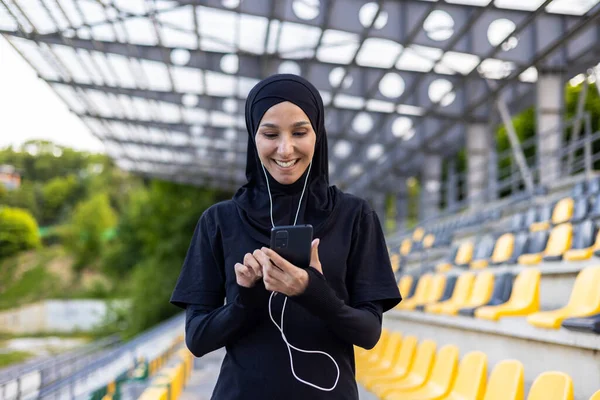 Muslim smiling lady in black hijab and sportswear listening to music in wired earphones while training at stadium. Active woman with phone adding new song to playlist for exercising on fresh air.