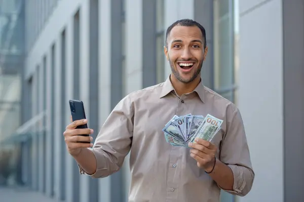 Adult hispanic man holding mobile phone and fan of dollar bills in hands with excited facial expression. Lucky guy in shirt boasting of winning cash in online lottery while standing outside.