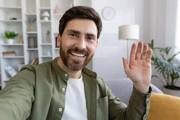 Adult male with stylish hairstyle waving hello with hand to camera while sitting on sofa over domestic interior background. Friendly guy in shirt greeting companion while starting new video chat.