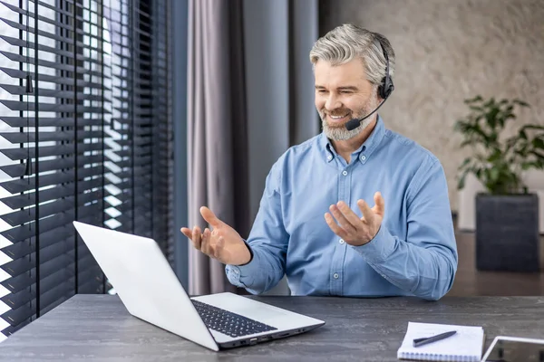 Elderly grey-haired man in denim shirt sitting at domestic workspace with laptop and having distant meeting with coworkers. Caucasian businessman in headset using online communication privileges.