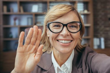 Gözlüklü, gülümseyen ve el sallayan, iyi donanımlı bir ofis ortamından uzaktan çalışan dost canlısı, olgun bir iş kadını..