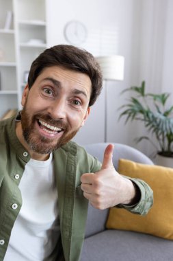 Sakallı, neşeli, olgun bir adam. Sıcak bir evde kanepede dinlenirken gülümsüyor ve güzel bir jest yapıyor..