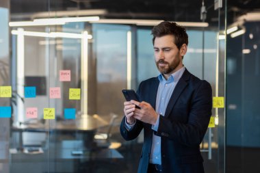 Modern ofiste akıllı telefon kullanan takım elbiseli ciddi bir işadamı. Finans, liderlik ve kurumsal yaşam tarzına odaklanmış.