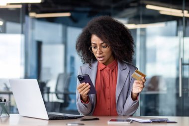 Gözlüklü, kıvırcık saçlı, akıllı telefon ve kredi kartıyla modern işyerinde çalışan profesyonel bir kadın..