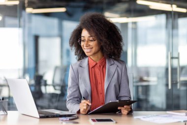 Kendine güvenen kıvırcık saçlı ve gözlüklü genç iş kadını ofis masasında bilgisayar ve belgelerle çalışıyor, profesyonelliği somutlaştırıyor ve odaklanıyor..