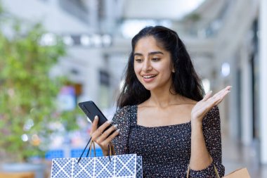 Neşeli Hintli kadın alışveriş merkezi ortamında renkli çantalar tutarken cep telefonu kullanıyor..