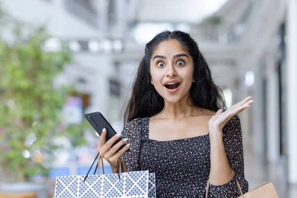 Surprised Indian female shopper with shopping bags and mobile phone in a mall, embodying the joy of retail therapy.