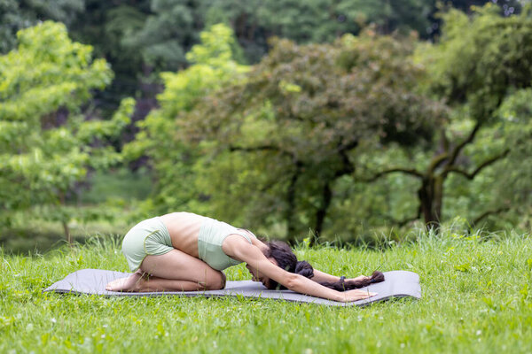 A young Indian woman in a tracksuit is deeply immersed in her yoga practice amidst the tranquil greenery of a lush park.