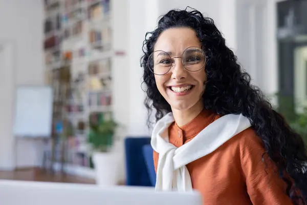 Side view of brunette female in round eyewear and white scarf over orange blouse smiling at camera. Friendly latina woman welcoming joining book club at college with satisfied facial expression.