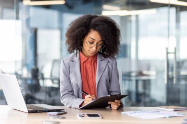 Kıvırcık saçlı profesyonel iş kadını, iş yerindeki ofisinde not defterine yazmaya odaklanıyor..
