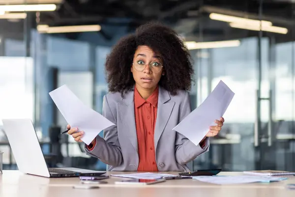 Young female professional in an office setting appears surprised, managing a high workload with a puzzled look and holding paperwork.