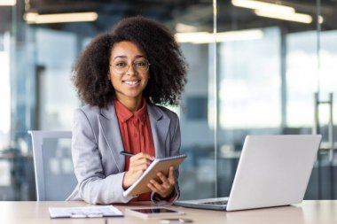 Beyaz yakalı bir işçi, bir dizüstü bilgisayar ve tabletle, mobilyalarla çevrili bir masada oturuyor. Kişisel bilgisayarında çalışırken gülümsüyor..