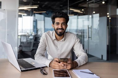 Hintli girişimci ofis masasında dizüstü bilgisayar, akıllı telefon ve not defteriyle çalışırken gülümsüyor..