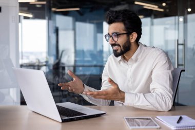 Online toplantı, konferans. Hintli genç adam, ofis çalışanı ve işadamı bilgisayarında video araması için dua ediyor, masada oturuyor, elleriyle işaret ediyor ve gülümsüyor..