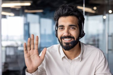 Yüzünde kulaklıkla gülümseyen Hintli bir işadamının portresi. Ofis ortamında el sallıyor..