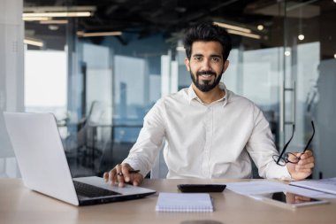 Beyaz yakalı bir işçi dizüstü bilgisayarla bir masada oturuyor ve gözlük takıyor, ofis ekipmanlarıyla bir mühendislik projesi üzerinde çalışıyor..