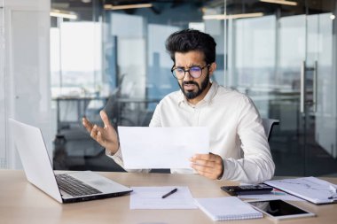 Beyaz yakalı bir işçi kişisel bilgisayar ve ofis malzemeleriyle bir masada oturuyor, bir dizüstü bilgisayar ve bir parça kağıt da dahil..