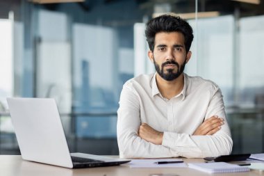 Sakallı bir adam bir masada oturuyor, kollarını kavuşturmuş, dizüstü bilgisayarın önünde..