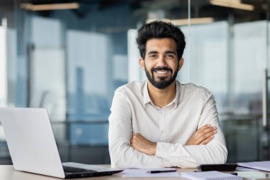 Profesyonel Hintli adam ofis masasında gülümsüyor, modern bir çalışma alanında dizüstü bilgisayarla çalışıyor, başarıyı ve güveni somutlaştırıyor..