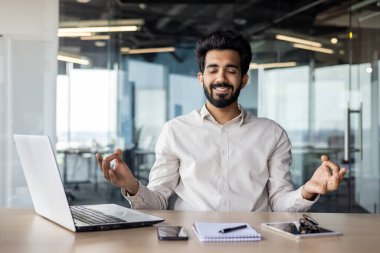 Beyaz yakalı bir işçi dizüstü bilgisayarla bir masada oturuyor. Etrafı binayla çevriliyken meditasyon yapıyor. Sakalı sakin bir ortam yaratıyor..