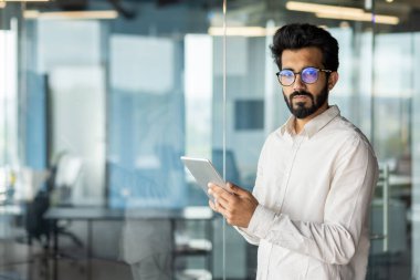 Ofiste sakallı ve bıyıklı beyaz yakalı bir işçi duruyor. Üzerinde kıyafet ve gözlük varken elinde tablet bir bilgisayar var..