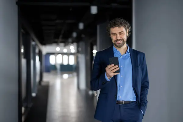 Mature experienced successful businessman in business suit walking inside office building, man walking down corridor with phone in hands, boss using app smartphone, reading and smiling contentedly