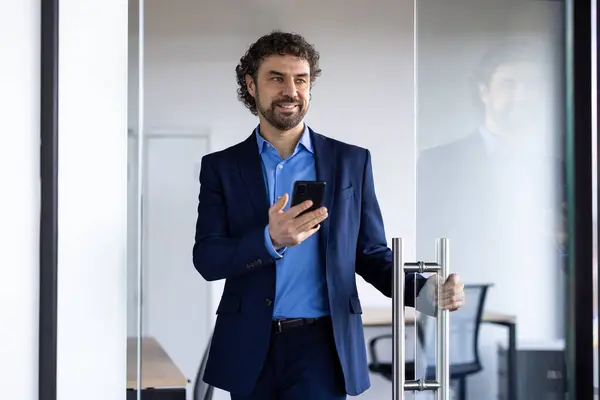 Successful joyful businessman opens glass office door, man with phone in hands smiling, satisfied with achievement results, boss in business suit.