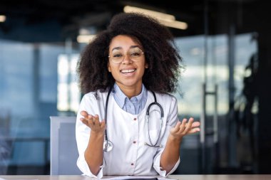 Başarılı, profesyonel üniformalı kadın cerrah dişlek gülümsemesiyle masa başında otururken el kaldırıyor. Pozitif siyah kadın iyi haberler ve tedavi sonuçlarının olumlu tahminlerini açıklamaya hazır..