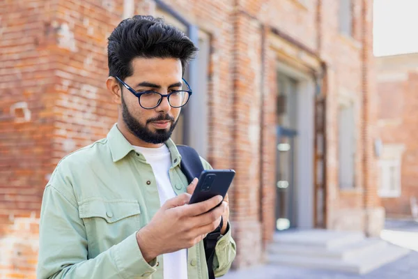 Young indian guy in eyeglasses looking at smartphone screen while standing with backpack outdoors. Focused young man checking online classes schedule for attending courses in university campus.