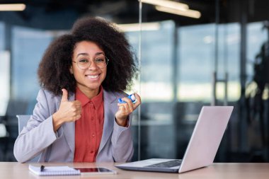 Takım elbiseli rahatlamış Latin bayan başparmak hareketi yapıyor ve dolapta otururken astım spreyi tutuyor. Gülümseyen yönetici bronşiyal hastalık saldırısından sonra kendini daha iyi hissediyor.