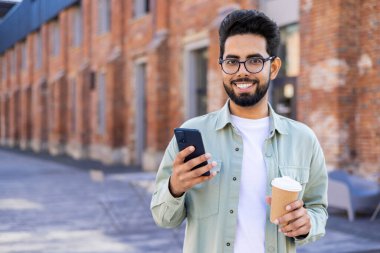 Gündüz yürürken elinde telefon ve plastik kahve fincanı tutan rahat bir Orta Doğulu adam. Esnek çalışma programı olan pozitif esmer serbest yazar içecek ve kaydırmalı yem için ara veriyor.