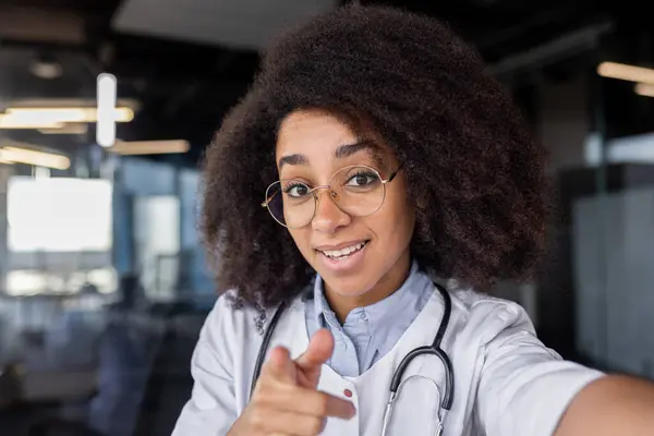 Diverse medic student in eyeglasses with bushy hairdo pointing at camera on blurred background. Pretty female intern recording video about importance of regular body checks for disease preventing.