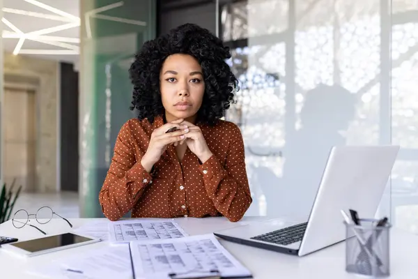 Serious diverse female boss strictly looking at camera while keeping chin on folded hands by table with clipboards. Millennial ceo putting computer aside and ready for discussing working moments.