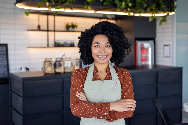 Satisfied black woman in apron standing with crossed arms on chest on background of bar counter. Confident small business owner in service sector ready for accepting clients in beginning of day.