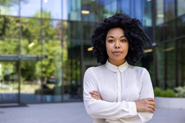 Serious black woman in white shirt standing with crossed arms on chest outside of glassy building with reflecting trees. Corporate middle age employer looking at camera with proud expression.