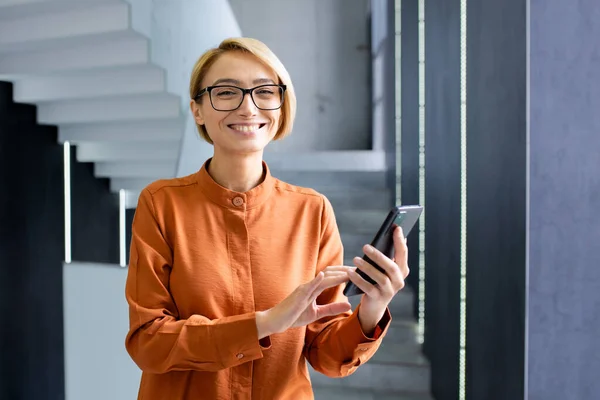 Cheerful caucasian lady in orange blouse holding mobile telephone while standing next to window in corporate building. Friendly female woman standing in hall and checking personal social media.