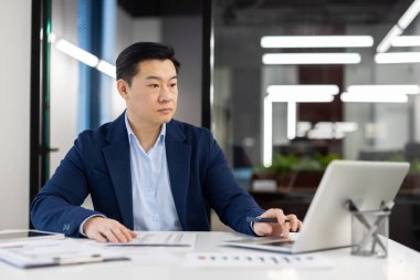 Ofis içinde kağıt işleri yapan ciddi düşünen bir adam. Masadaki verileri dolduran, kalemle yazan, iş yerinde dizüstü bilgisayarı olan bir iş adamı..