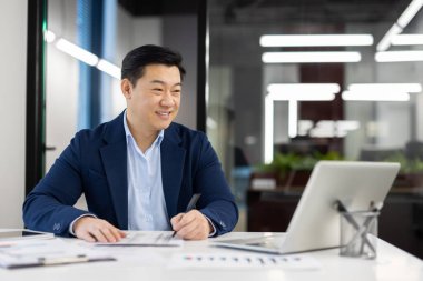 İş yerindeki başarılı erkek finansör muhasebeciden memnun kalan Asyalı işadamı, hesap sözleşmeleri ve raporlarla çalışmaktan mutlu bir şekilde gülümsüyor..