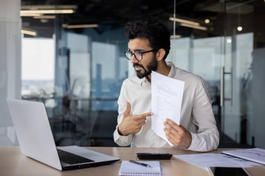 Ciddi genç Hintli erkek işadamı internette toplantı yapıyor, ofiste çalışıyor, masada oturuyor, bilgisayardaki bir video görüşmesinde konuşuyor ve kameraya belgeleri gösteriyor..