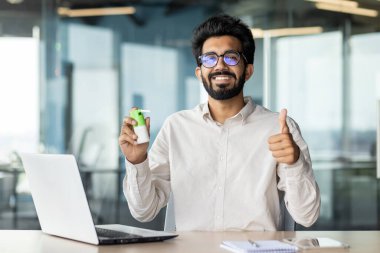 Ofisteki bir masada oturmuş elinde boğaz spreyi tutan ve parmaklarıyla süper bir jest yapan gülümseyen Hintli bir adamın portresi..