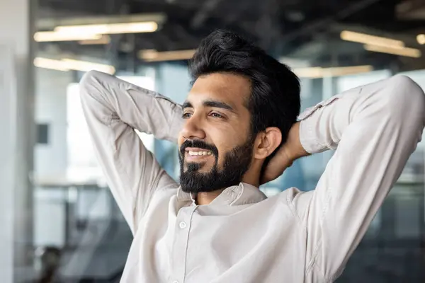 Happy and successful young Indian man, businessman and office worker sitting at workplace with hands behind head and looking ahead. Resting, satisfied with the work and the result. Close-up photo.